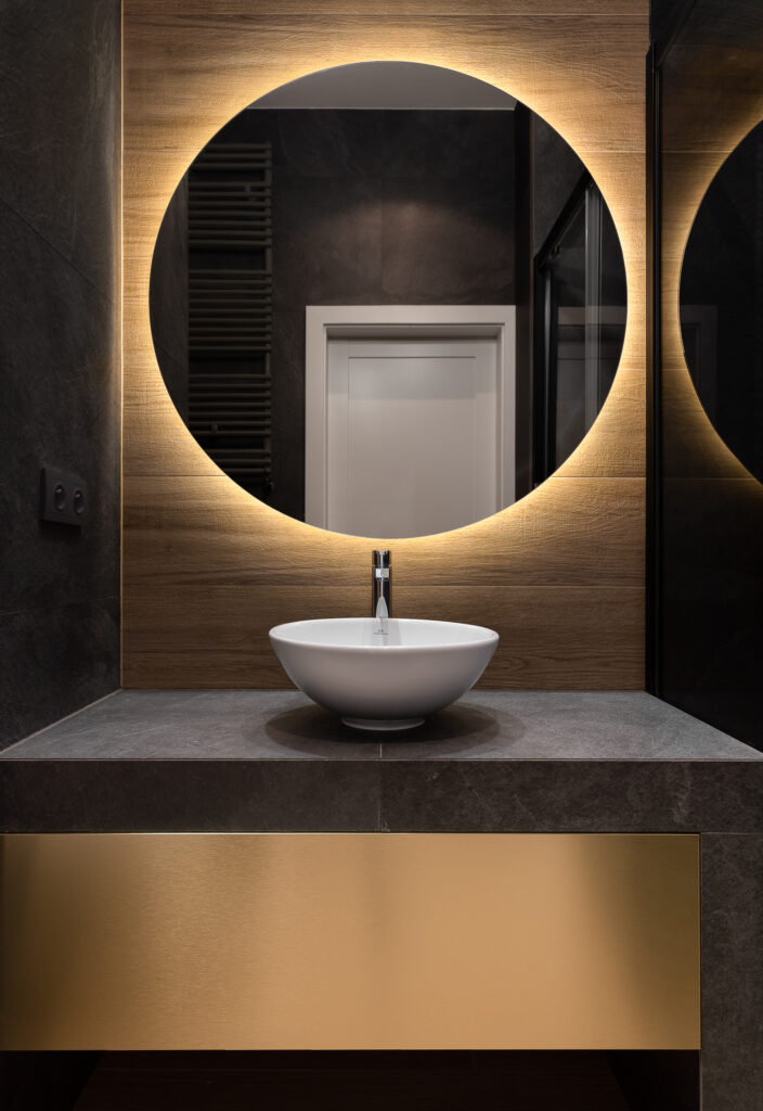 Gwiazdą tego projektu łazienki jest okrągłe, podświetlone lustro, którego poświata rzucona na drewniane panele nawiązuje kolorystycznie do złotej szuflady.